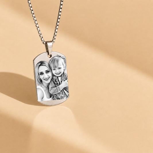 A silver dog tag necklace with a custom black-and-white photo of a woman and child, hanging from a silver ball chain against a warm beige background with soft shadows.