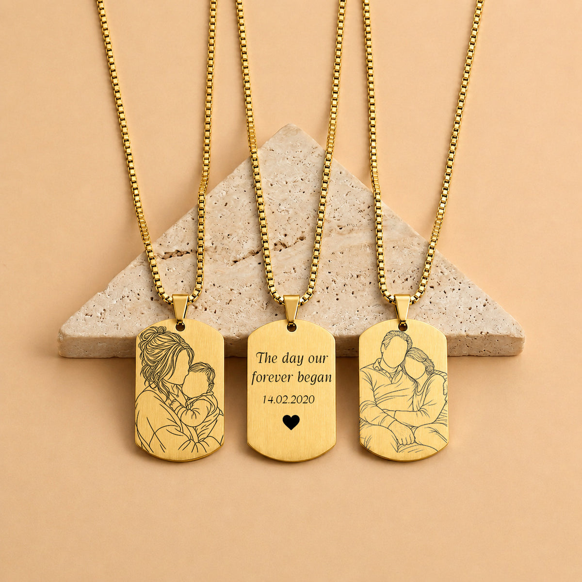 Three gold-toned dog tag necklaces with ball chains are displayed on a beige stone pedestal against a warm peach background. The left and right tags feature engraved couple portraits, while the center tag has a simple engraving reading 'The day our forever began 14.