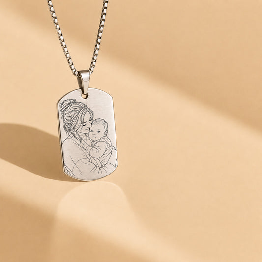 A silver dog tag necklace with an engraved portrait of a mother holding her baby, suspended from a silver ball chain against a warm beige background with soft shadows.
