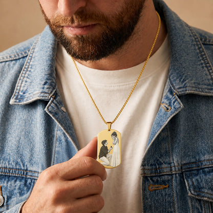 A close-up of a man wearing a gold dog tag necklace with an illustration of a kneeling man and a woman in a white dress. He is wearing a white t-shirt and a blue denim jacket against a warm beige background.