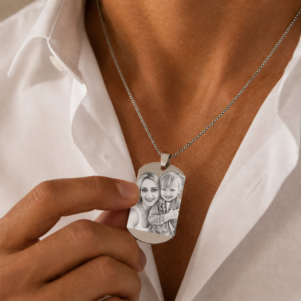 A close-up of a person wearing a silver chain necklace with a custom engraved dog tag pendant. The pendant features a black-and-white sketch of a woman and child, held by a hand against the person's white shirt.