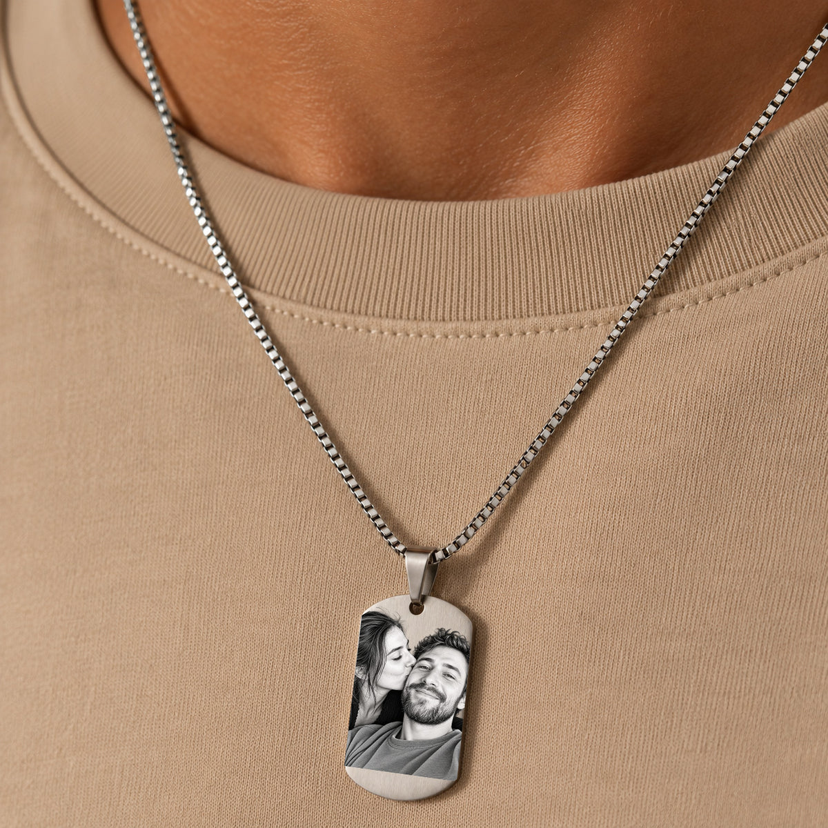 A close-up of a person wearing a silver chain necklace with a custom photo printed on a rectangular dog tag pendant, set against a beige crewneck shirt.