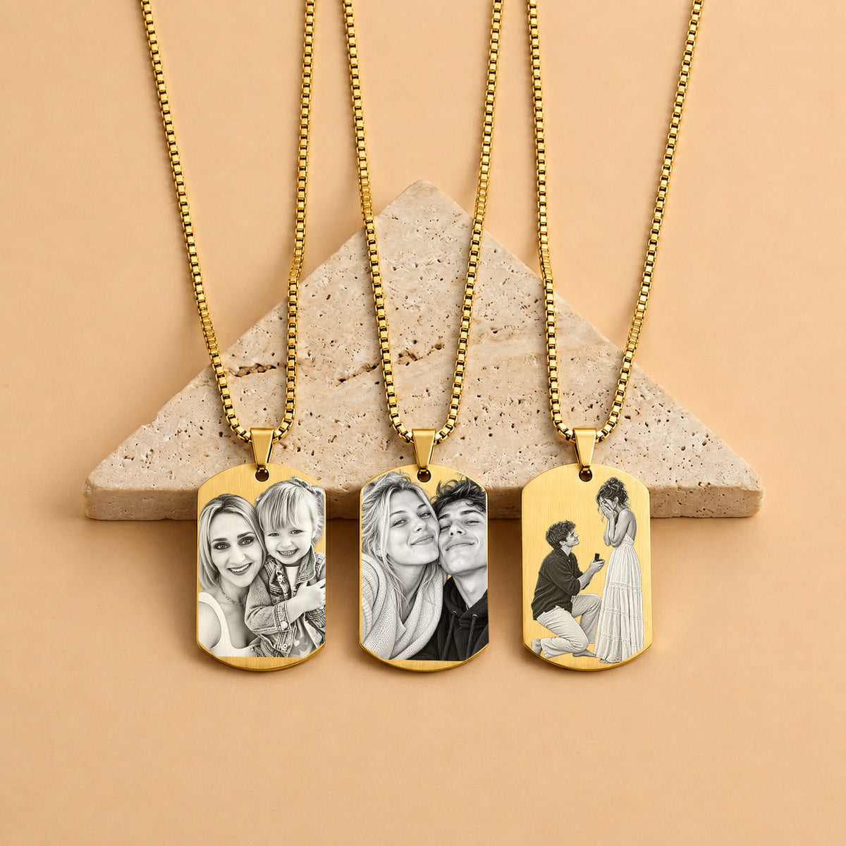 Three gold-tone custom photo dog tag necklaces with ball chains are displayed on a beige stone block against a warm peach background. Each pendant shows a different custom couple or family portrait illustration.